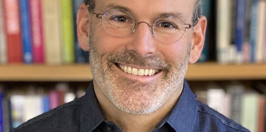 Smiling man with glasses wearing a dark shirt, standing in front of a bookshelf filled with colorful books.